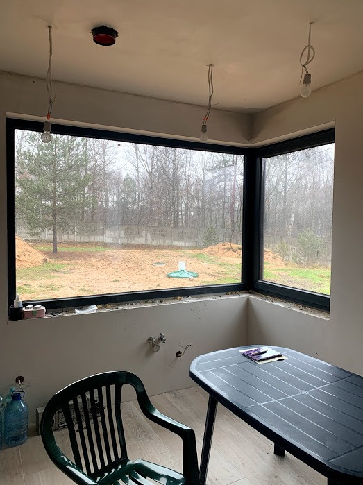 Narożne okno z czarną ramą w surowym wnętrzu, widok na ogród z drzewami. Stolik i krzesło w pomieszczeniu. Niedokończone wykończenie ścian.