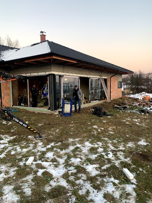 Montaż przesuwnych szklanych drzwi tarasowych w surowym stanie budynku. Widoczny pracownik z podnośnikiem, drabina i ekipa montażowa wewnątrz.