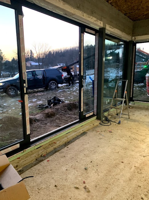 Montaż przesuwnych okien z czarnymi ramami. Na zewnątrz widoczny samochód z dźwigiem podczas instalacji. Wnętrze w stanie surowym.