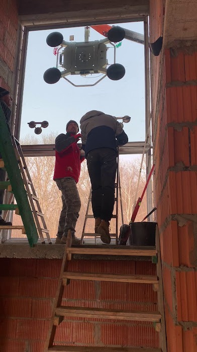 Montaż dużego okna przez dwóch monterów za pomocą przyssawki próżniowej w surowym stanie budynku z cegły ceramicznej. Widoczne drabiny i narzędzia.