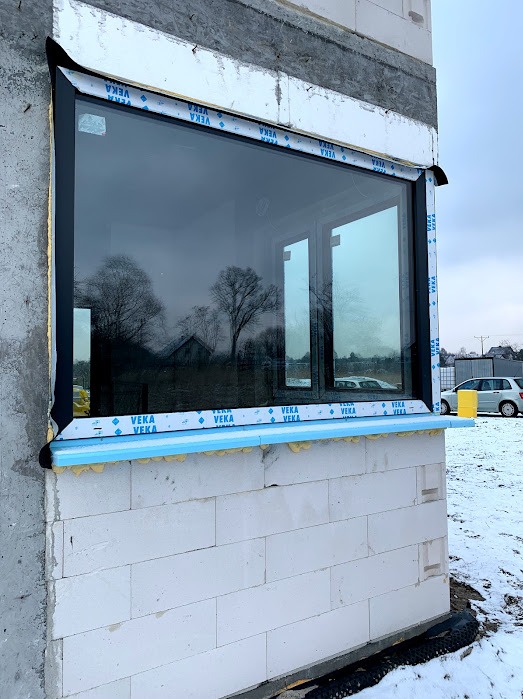 Czarne okno Veka w surowej ścianie z betonu komórkowego; widoczne uszczelnienia i taśmy montażowe. W szybie odbija się krajobraz.