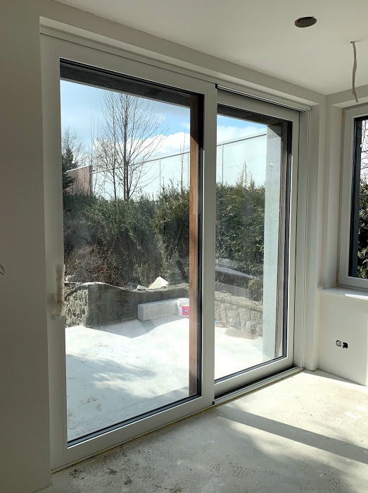 Nowoczesne, przesuwne okno tarasowe z białymi ramami, widok na ogród. Elementy wykończeniowe w kolorze drewna. Jasne wnętrze w trakcie prac wykończeniowych.