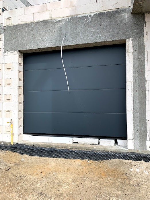 Szara segmentowa brama garażowa w surowym otworze budynku z białego bloczka, widoczny przewód instalacyjny i żółty zawór gazowy.