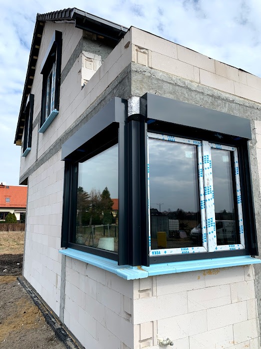 Narożne okno z roletami zewnętrznymi w surowym budynku, widoczne nieotynkowane ściany z betonu komórkowego i fragmenty innych okien na wyższych kondygnacjach.