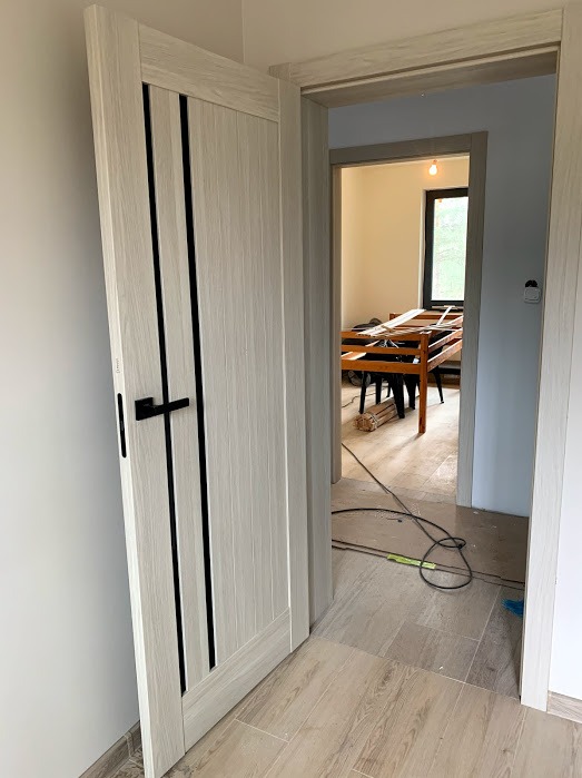 Otwarta, jasna dębowa drzwi wewnętrzne z czarnymi, pionowymi wstawkami i klamką, widok na wnętrze z drewnianym stołem i oknem. Podłoga wyłożona płytkami imitującymi drewno.