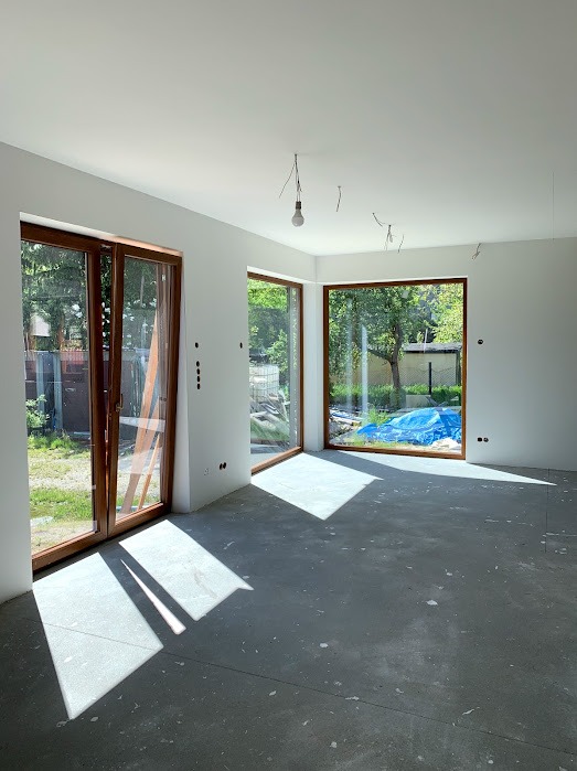Wnętrze w stanie deweloperskim z dużymi oknami drewnianymi i przesuwnymi drzwiami balkonowymi. Widoczne przygotowania do wykończenia, surowy beton na podłodze.
