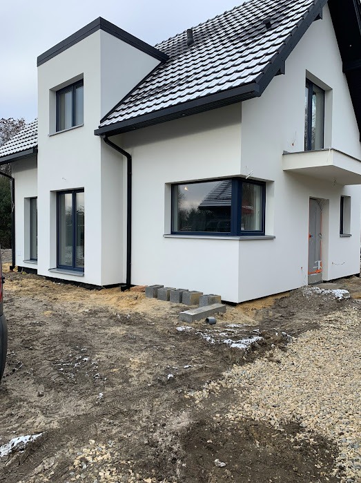 Nowoczesny dom jednorodzinny z ciemnymi oknami na tle ziemi i drobnego gruzu. Częściowo ośnieżony dach z ciemną dachówką. Widoczne bloczki betonowe przy ścianie.