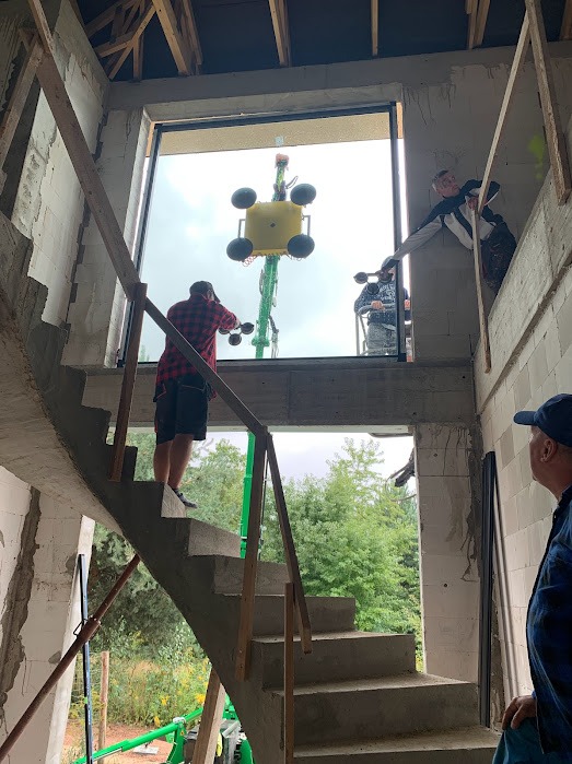 Montaż okna dźwigiem z przyssawkami w surowym budynku. Widoczni pracownicy na różnych poziomach, schody betonowe i zieleń za oknem. Etap budowy.