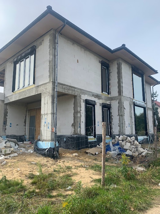 Surowy stan dwukondygnacyjnego domu z zamontowanymi oknami z roletami zewnętrznymi, widok z zewnątrz, teren budowy z gruzem i roślinnością.