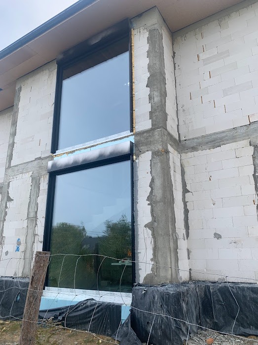 Nowe okna z czarnymi ramami zamontowane w surowym budynku z białego bloczka, widoczne ocieplenie i przymocowana siatka zabezpieczająca.