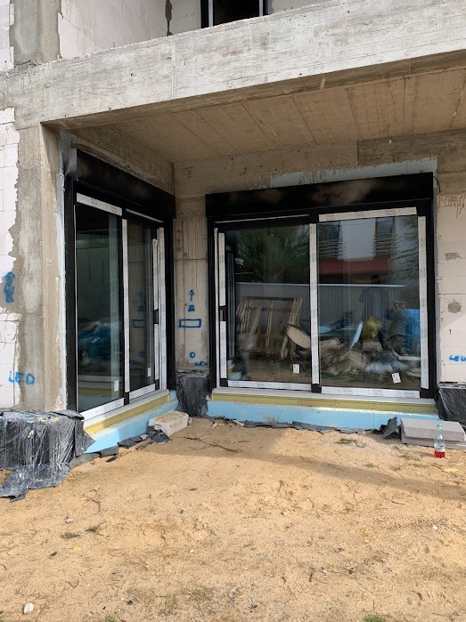 Dwa okna przesuwne z czarnymi roletami w surowym stanie budynku. Widoczne izolacje i piasek na zewnątrz. Okna z białymi ramami.