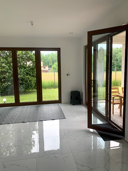 Nowoczesne, brązowe drzwi balkonowe otwarte na taras. Widok na ogród przez duże okno. Białe marmurowe płytki podłogowe. W tle zielony taboret.