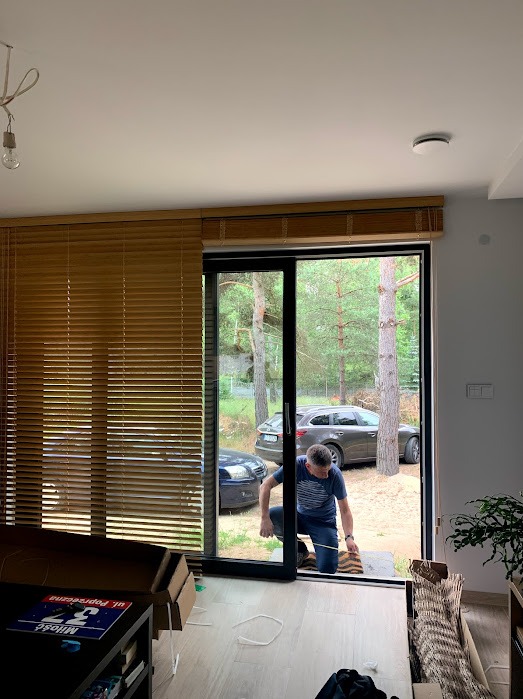 Montaż drewnianych żaluzji w oknie przesuwnym. Mężczyzna w trakcie pracy mierzy odległość. Widok na zewnątrz, gdzie widać zieleń i samochody.