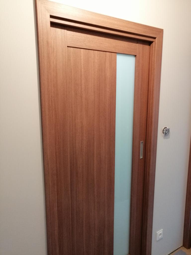 Zamknięte, przesuwne drzwi wewnętrzne z okleiną drewnopodobną i pionowym, matowym przeszkleniem, osadzone w ciemnobrązowej ościeżnicy.