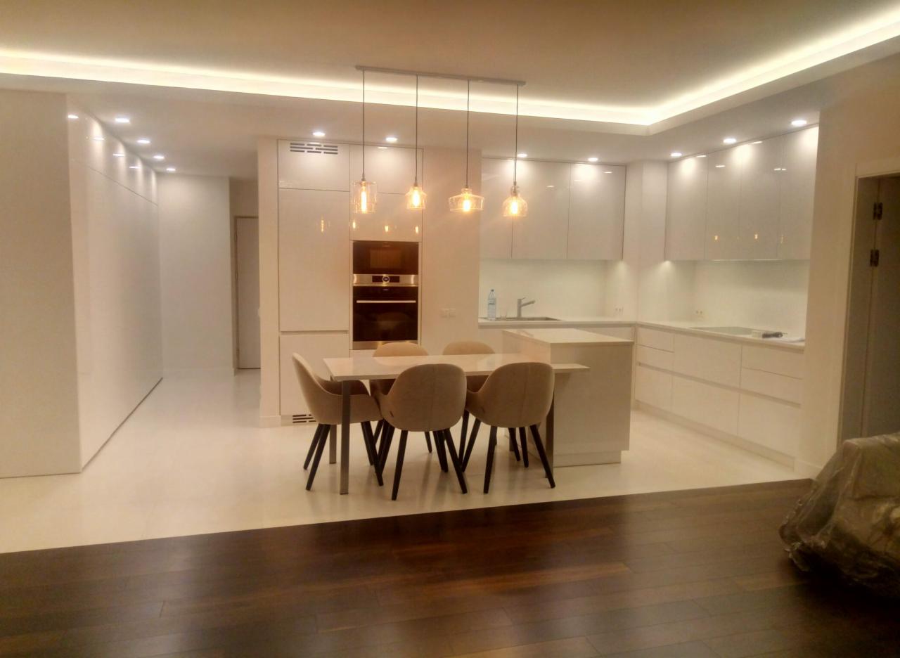 Nowoczesna, jasna kuchnia z białymi, błyszczącymi szafkami, okrągłym piekarnikiem w zabudowie, marmurowym blatem stołu i beżowymi krzesłami z ciemnymi nogami, oświetlona wiszącymi lampami...