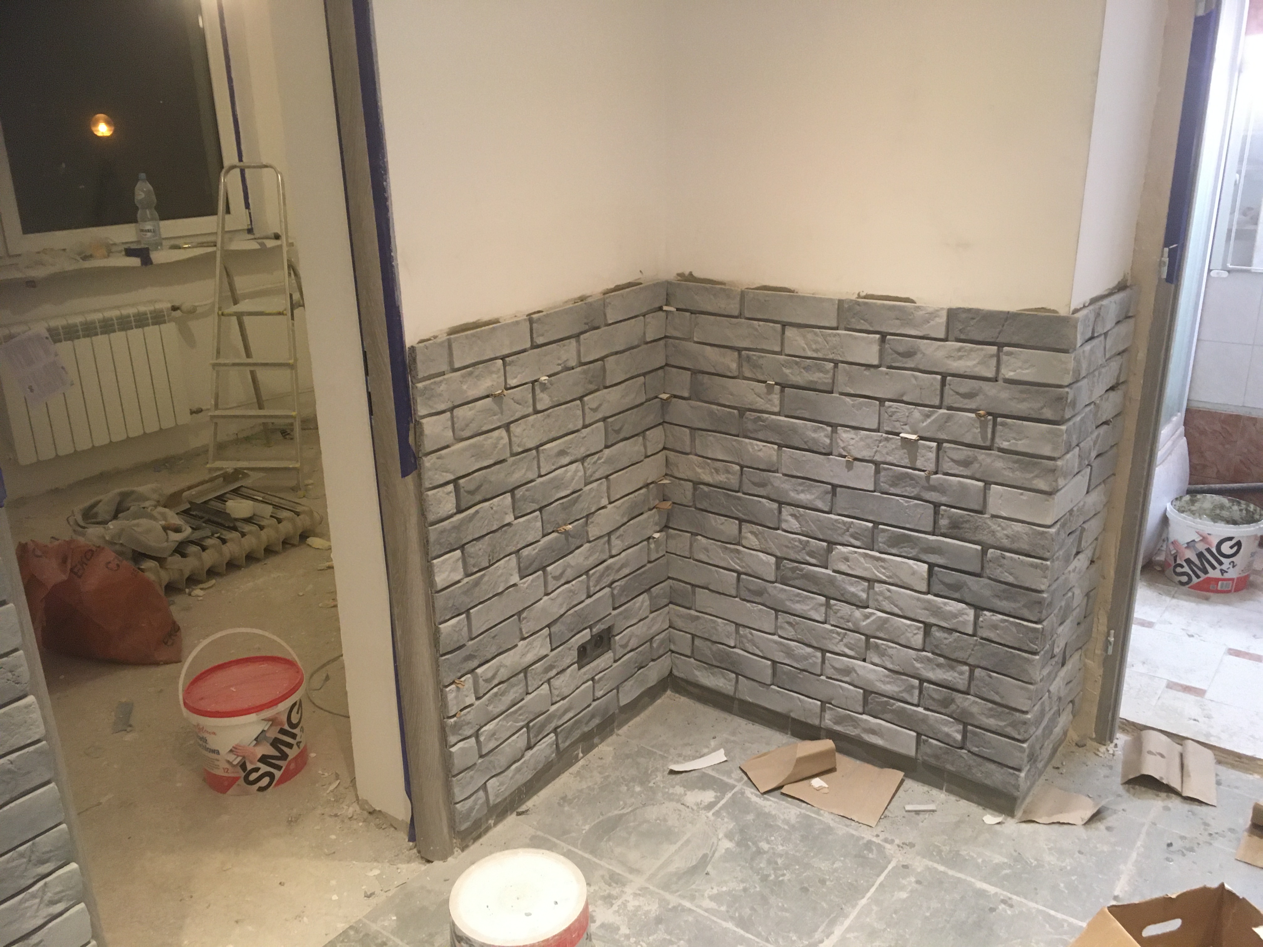 Fragment ściany z szarej cegły dekoracyjnej, widoczne puszki z zaprawą, drabina w tle, etap prac wykończeniowych.