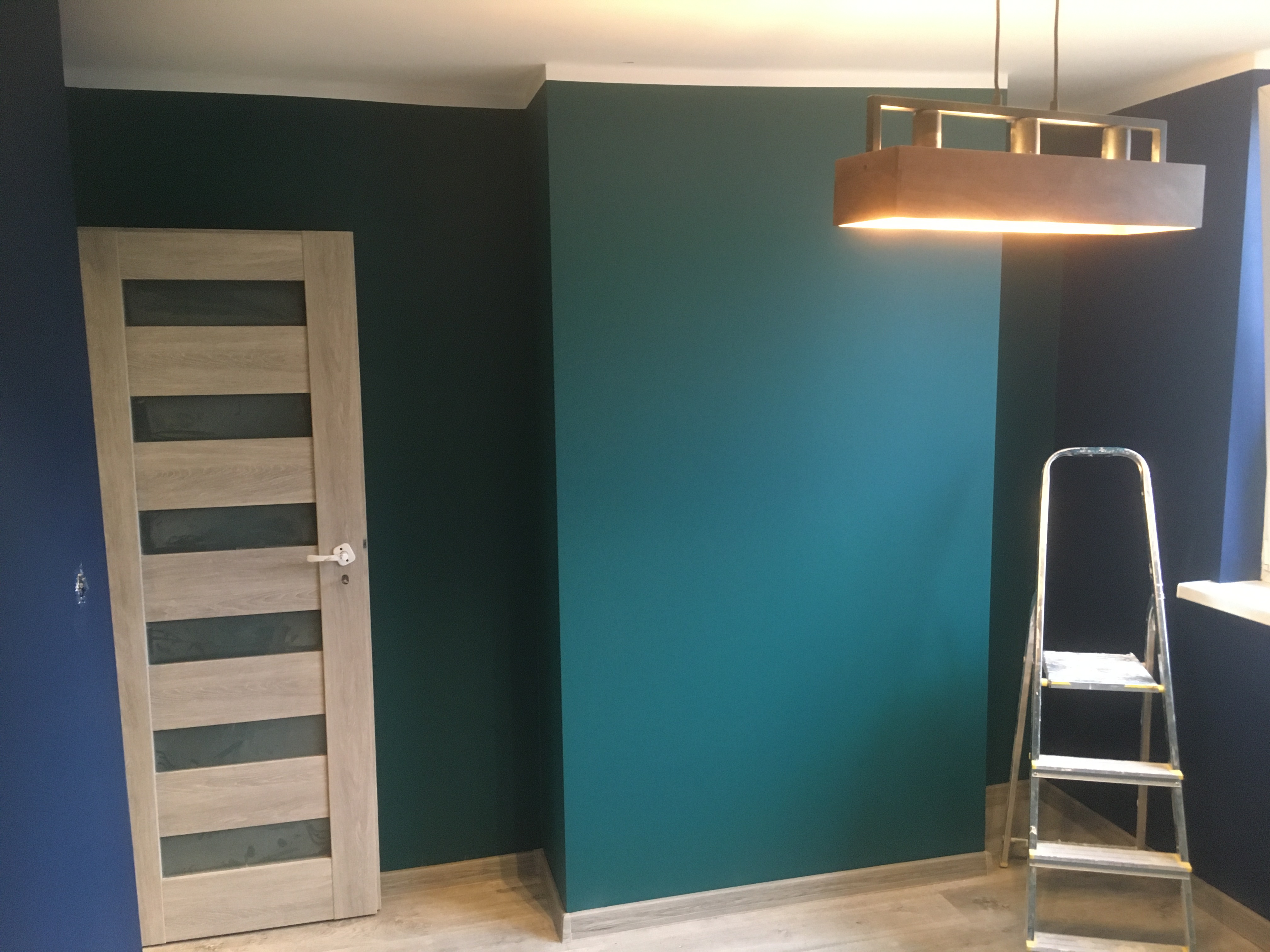 Świeżo pomalowane turkusowe ściany w pokoju z drewnianą podłogą, widoczna drabina malarska, nowoczesna lampa sufitowa i drzwi z przeszkleniami.