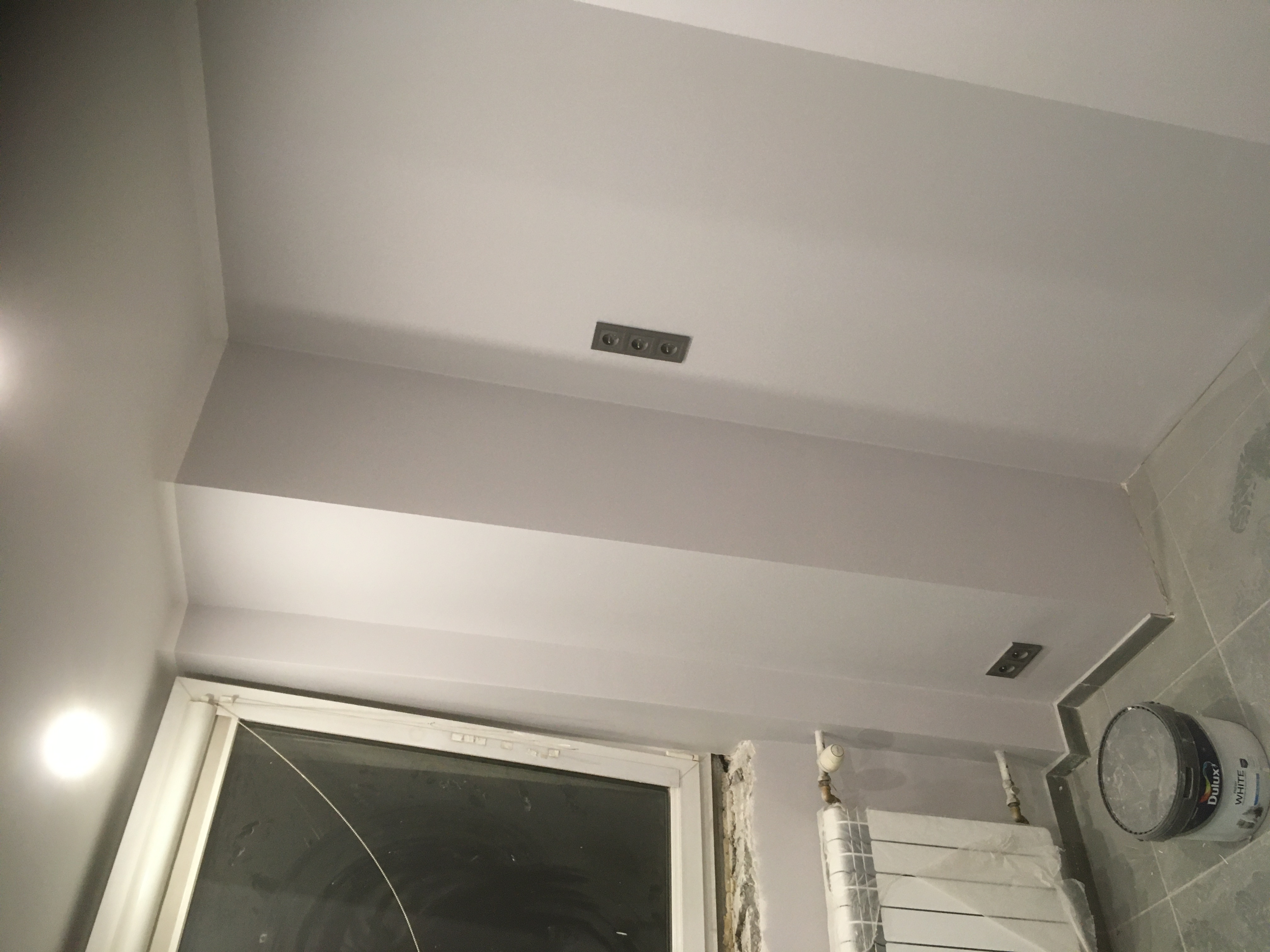 Świeżo pomalowane na szaro wnętrze łazienki w trakcie remontu, widoczna puszka farby Dulux White, grzejnik owinięty folią, gniazdka elektryczne w ścianie, okno z uszkodzoną szybą i fragment ściany...