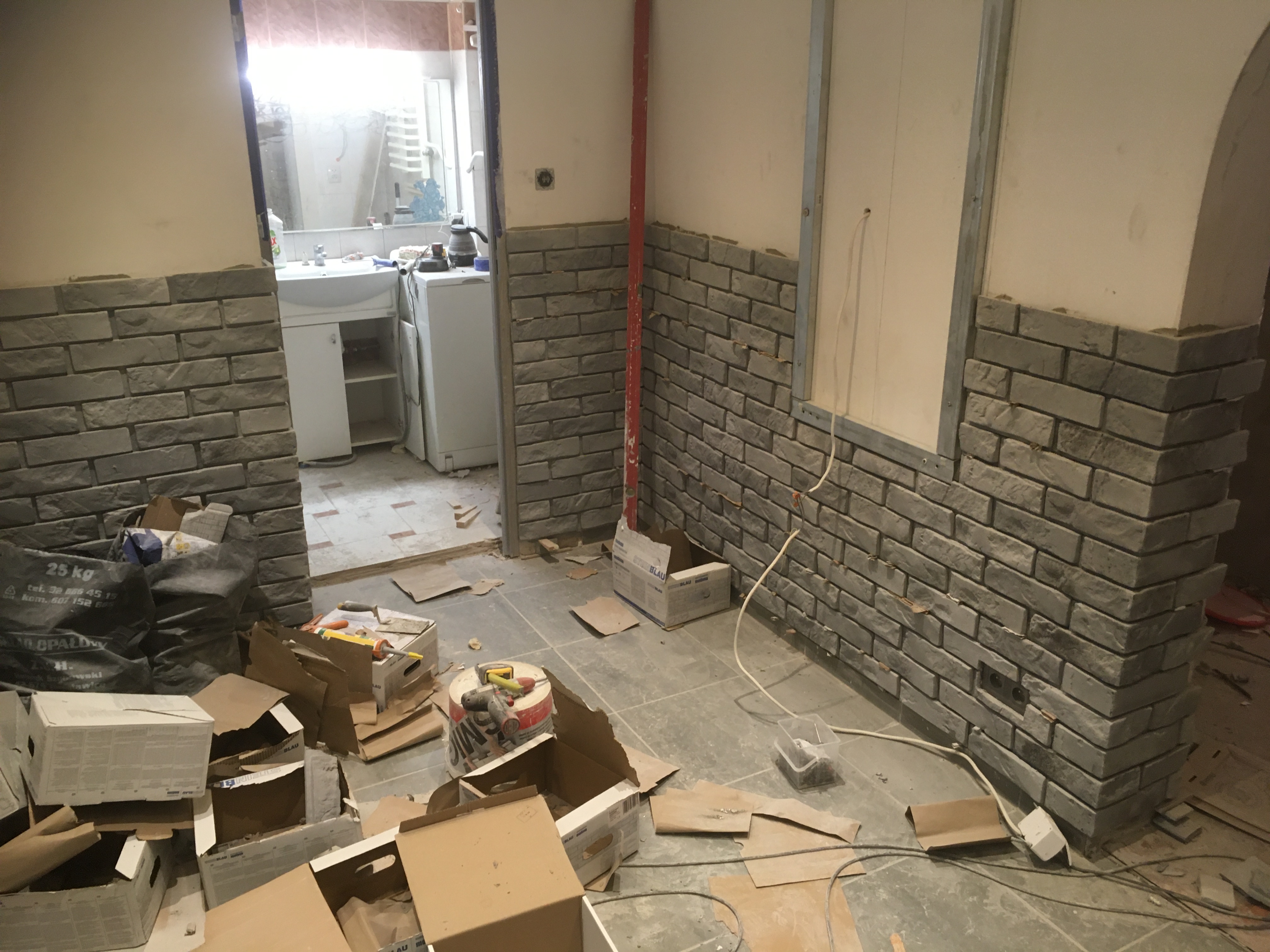 Wnętrze w trakcie remontu z częściowo wykończonymi ścianami z szarej cegły dekoracyjnej, widoczne narzędzia, materiały budowlane oraz niezabezpieczone kable elektryczne, w tle umywalka i pralka...