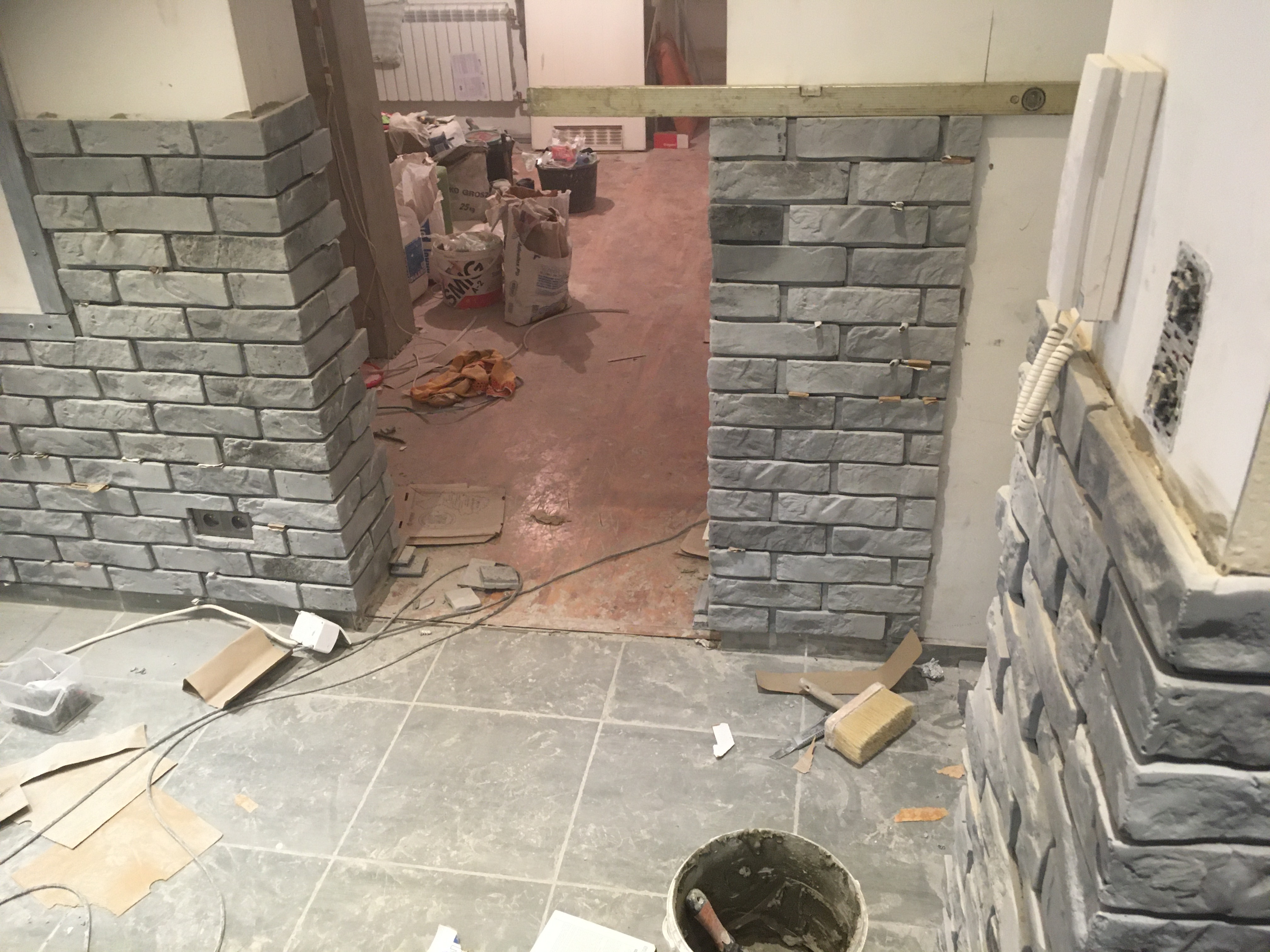 Montaż dekoracyjnej ściany z imitacji cegły wewnątrz pomieszczenia, widoczne narzędzia i materiały budowlane, poziomica na ścianie, surowy stan wykończenia.