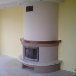 Otynkowany kominek z brązowym blatem i marmurową podstawą, w jasnym pomieszczeniu z żółtą ścianą.