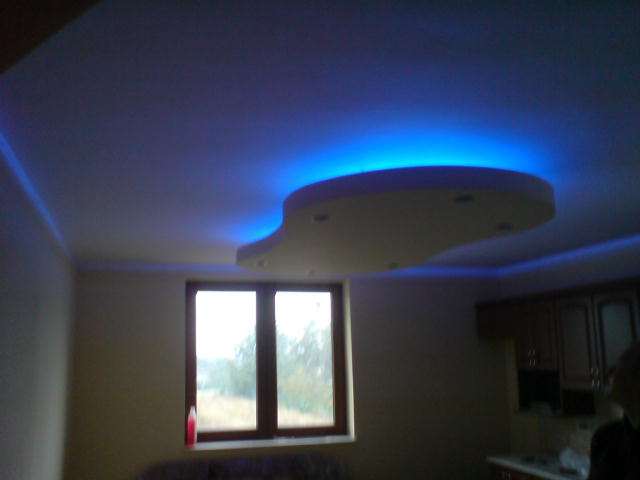 Wykończone wnętrze z podwieszanym sufitem o nieregularnym kształcie, podświetlonym intensywnym niebieskim światłem LED, widoczne okno i fragment zabudowy kuchennej.