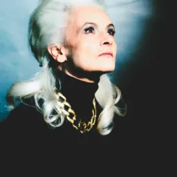 Portret kobiety o siwych włosach, w czarnym golfie i złotym łańcuchu, spoglądającej w górę na tle niebieskiego oświetlenia. Ujęcie od dołu, podkreślające wyraz twarzy.