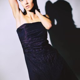 Fotografia niekonwencjonalna Ilona Bałon - Portret kobiety w fioletowej sukni bez ramiączek i kapeluszu, pozującej na jasnym tle z wyraźnym cieniem. Ujęcie studyjne z mocnym światłem podkreślającym rysy twarzy.