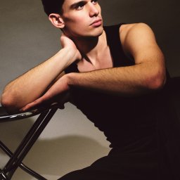Fotografia niekonwencjonalna Ilona Bałon - Mężczyzna w czarnym podkoszulku opiera się o metalowe krzesło, spoglądając w bok. Portret w stonowanej kolorystyce, z delikatnym oświetleniem.