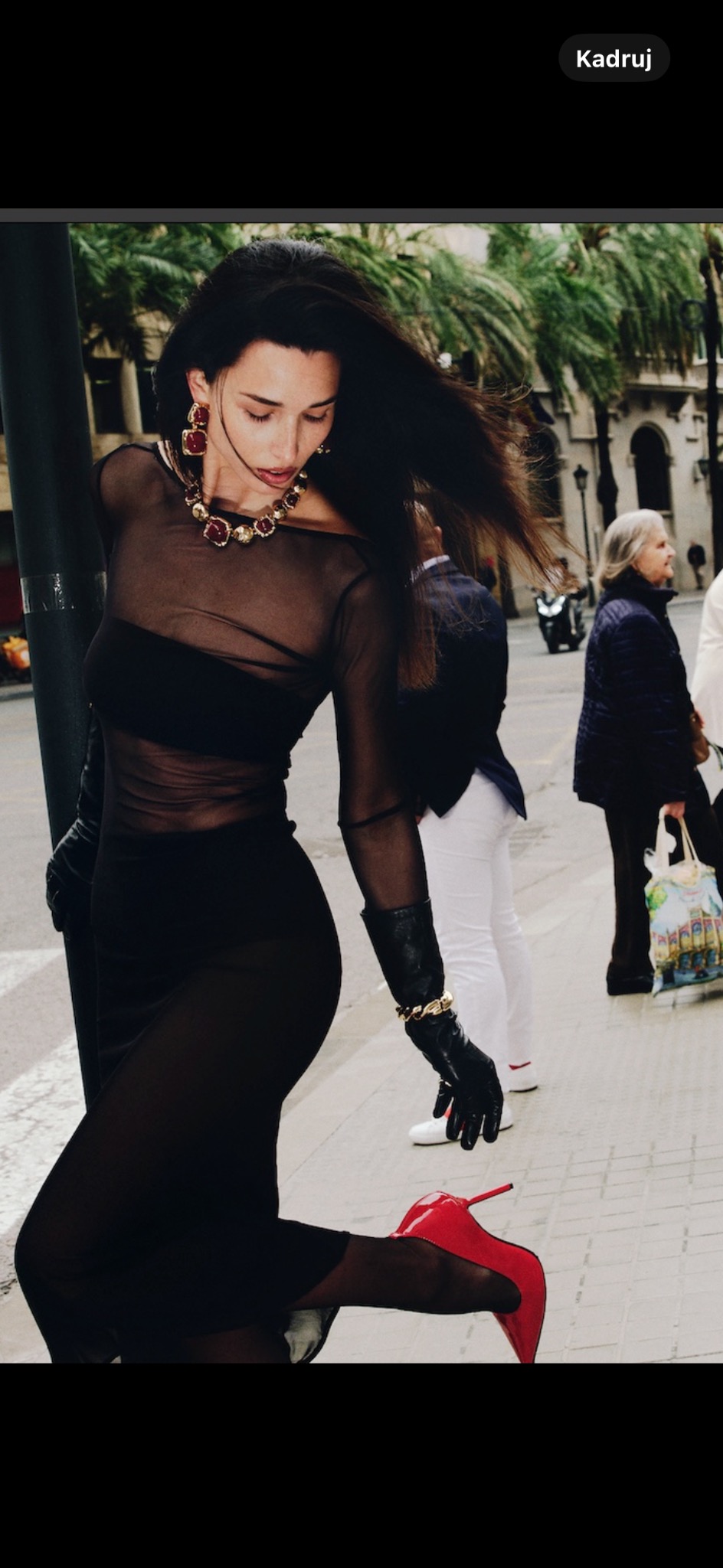 Dynamiczne ujęcie modelki w czarnej, prześwitującej sukience i czerwonych szpilkach na tle miejskiej scenerii z palmami, uchwycone w ruchu.