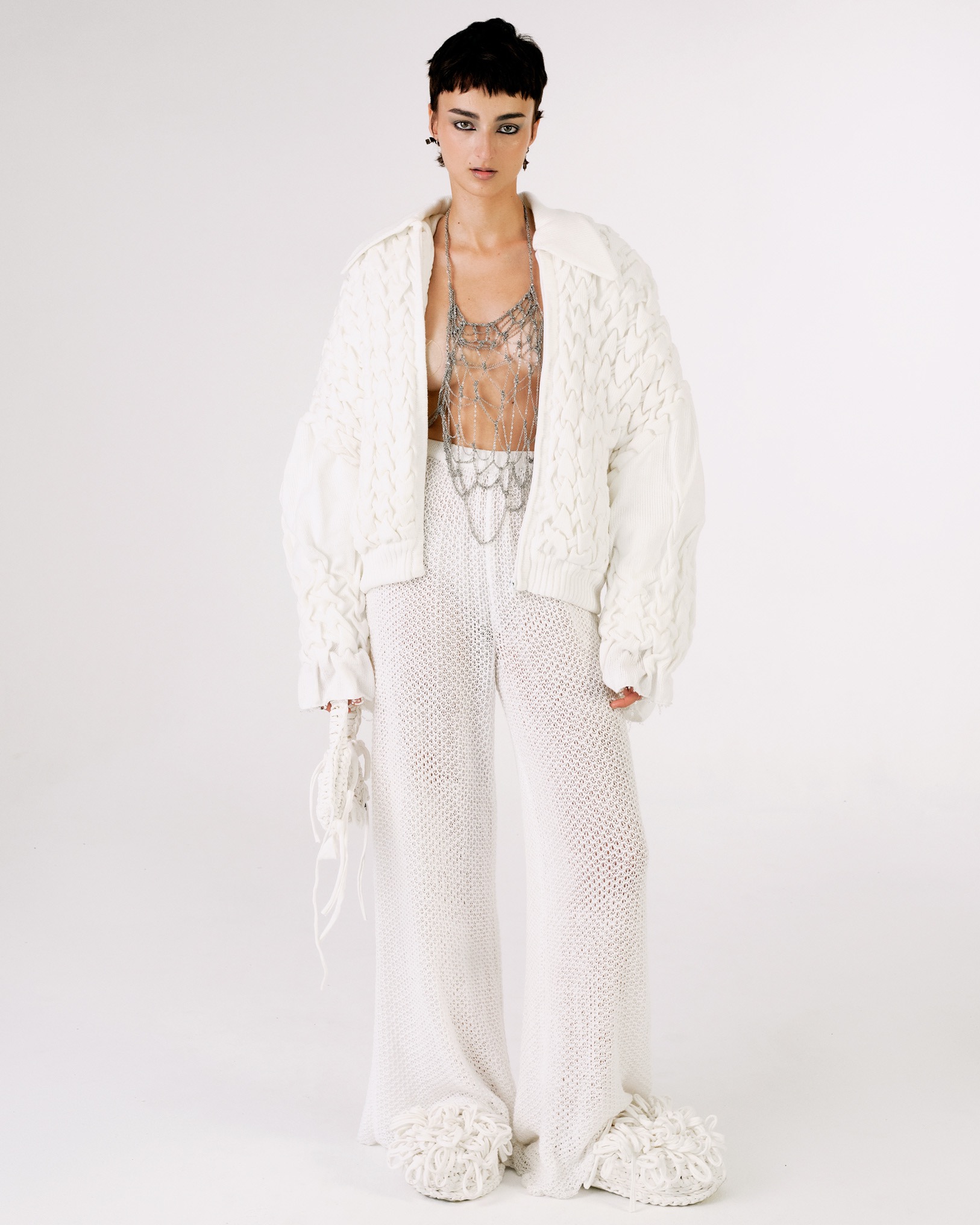 Modelka w białym, ażurowym stroju z grubym swetrem i srebrną ozdobą na ciele, na białym tle. Uwagę przykuwają nietypowe buty z frędzlami.