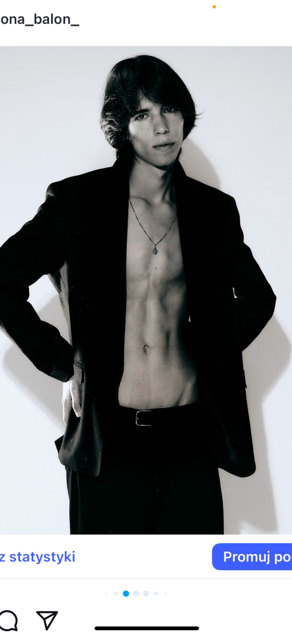 Czarno-biały portret mężczyzny w marynarce, bez koszuli, z łańcuszkiem, pozującego na jasnym tle. Minimalistyczny styl, ostre światło.