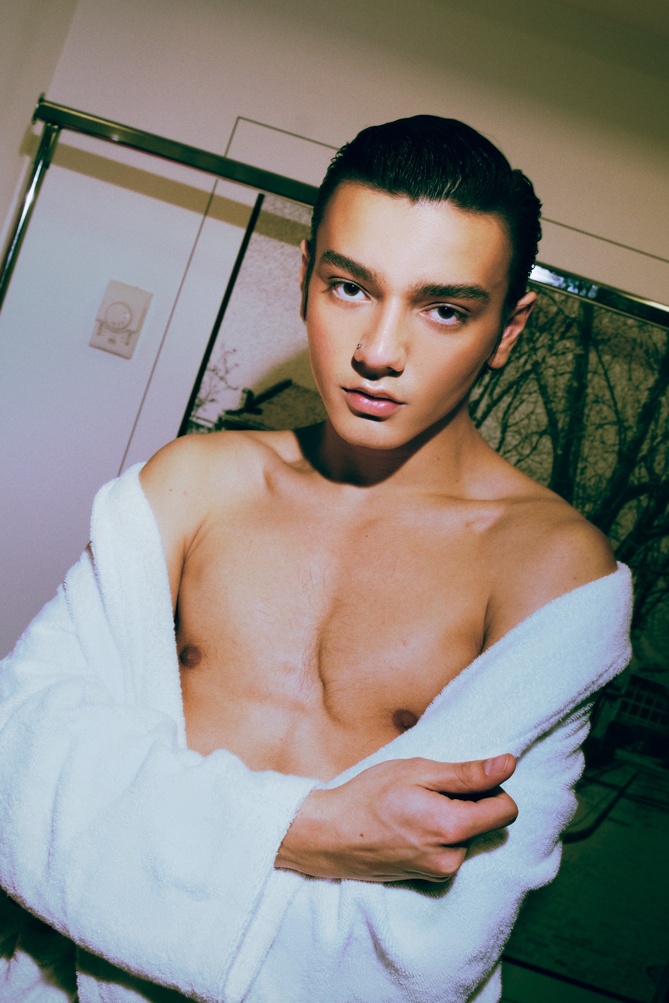 Mężczyzna w białym szlafroku, z piercingiem w nosie, pozuje na tle białej ściany i grafiki z motywem drzew. Zdjęcie oświetlone z góry, z widoczną klatką piersiową.