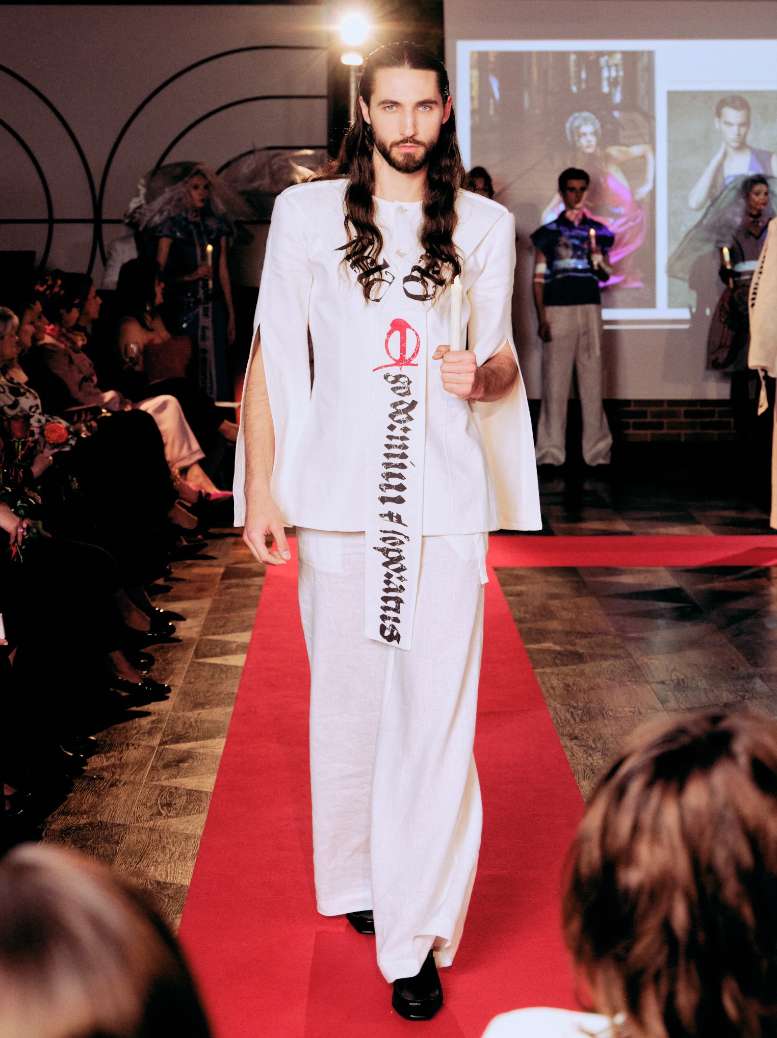Mężczyzna w białym stroju prezentuje kreację na pokazie mody w Niżnej Łące. Czerwony dywan, publiczność w tle, oświetlenie sceniczne.