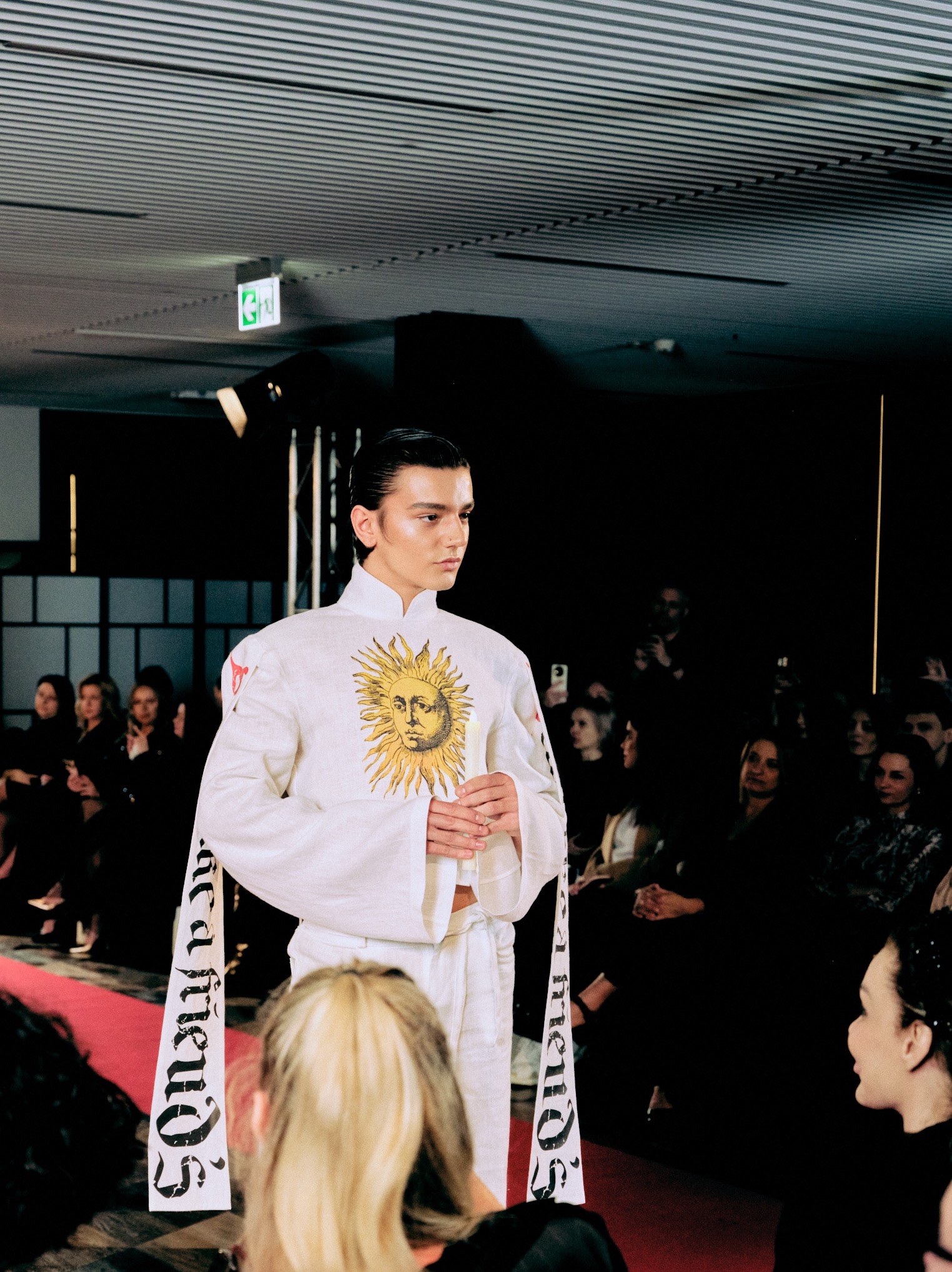 Model w białym stroju z motywem słońca prezentuje się na wybiegu podczas pokazu mody. Widoczna publiczność i oświetlenie sceniczne.