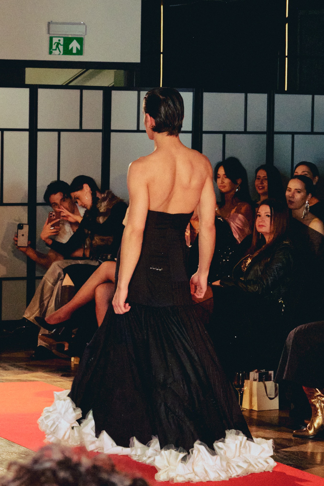 Model w czarnej sukni z falbanami prezentuje się na wybiegu podczas pokazu mody. Widzowie obserwują i robią zdjęcia telefonami. Czerwony dywan, eleganckie wnętrze.
