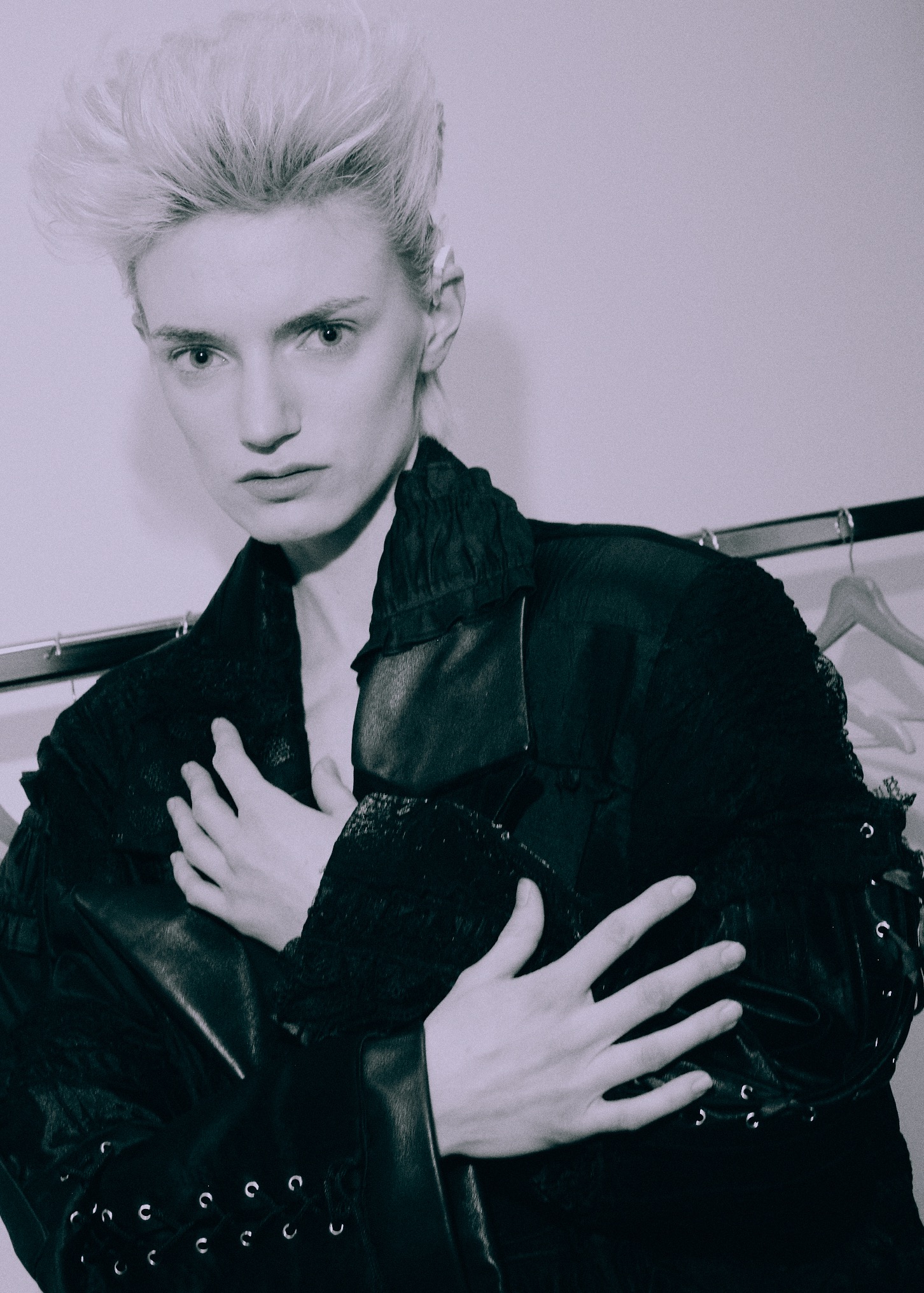 Portret osoby o jasnych włosach w czarnej, teksturowanej kurtce. Ujęcie w szarościach, z delikatnym oświetleniem i garderobą w tle. Charakterystyczny makijaż i stylizacja.