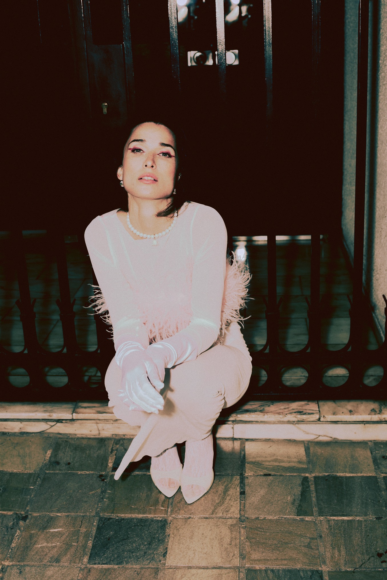 Kobieta w pastelowym stroju i rękawiczkach pozuje na tle metalowej bramy. Uwagę przykuwa jej makijaż z różowym akcentem oraz perłowy naszyjnik.