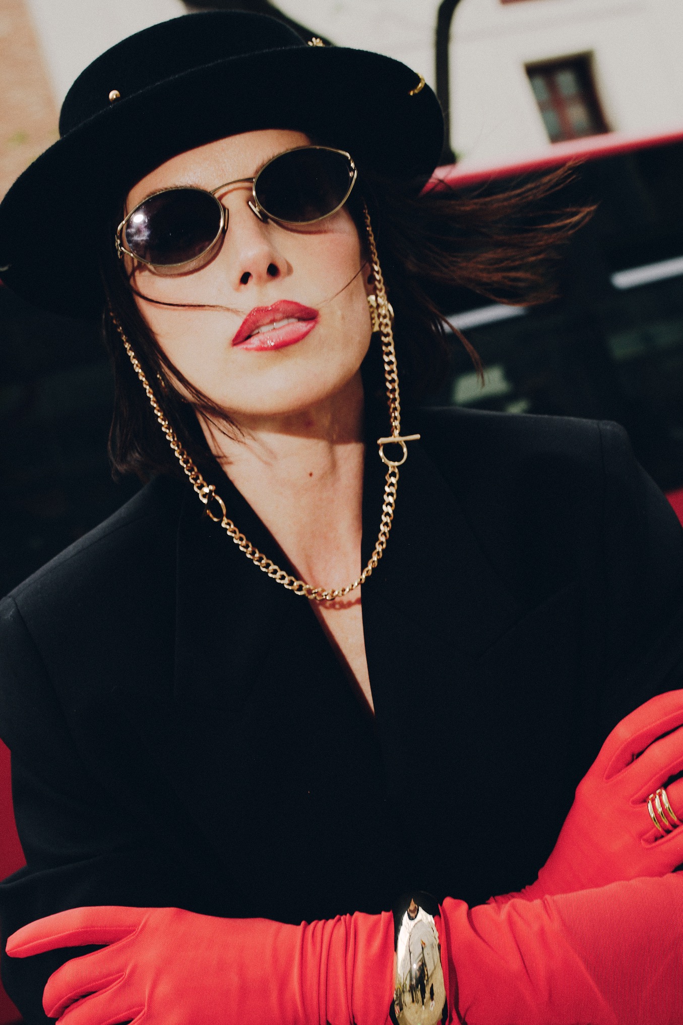 Stylowa kobieta w czarnym kapeluszu i okularach, z czerwoną szminką i rękawiczkami, pozuje na tle miejskiego krajobrazu. Złote łańcuszki dodają jej szyku.