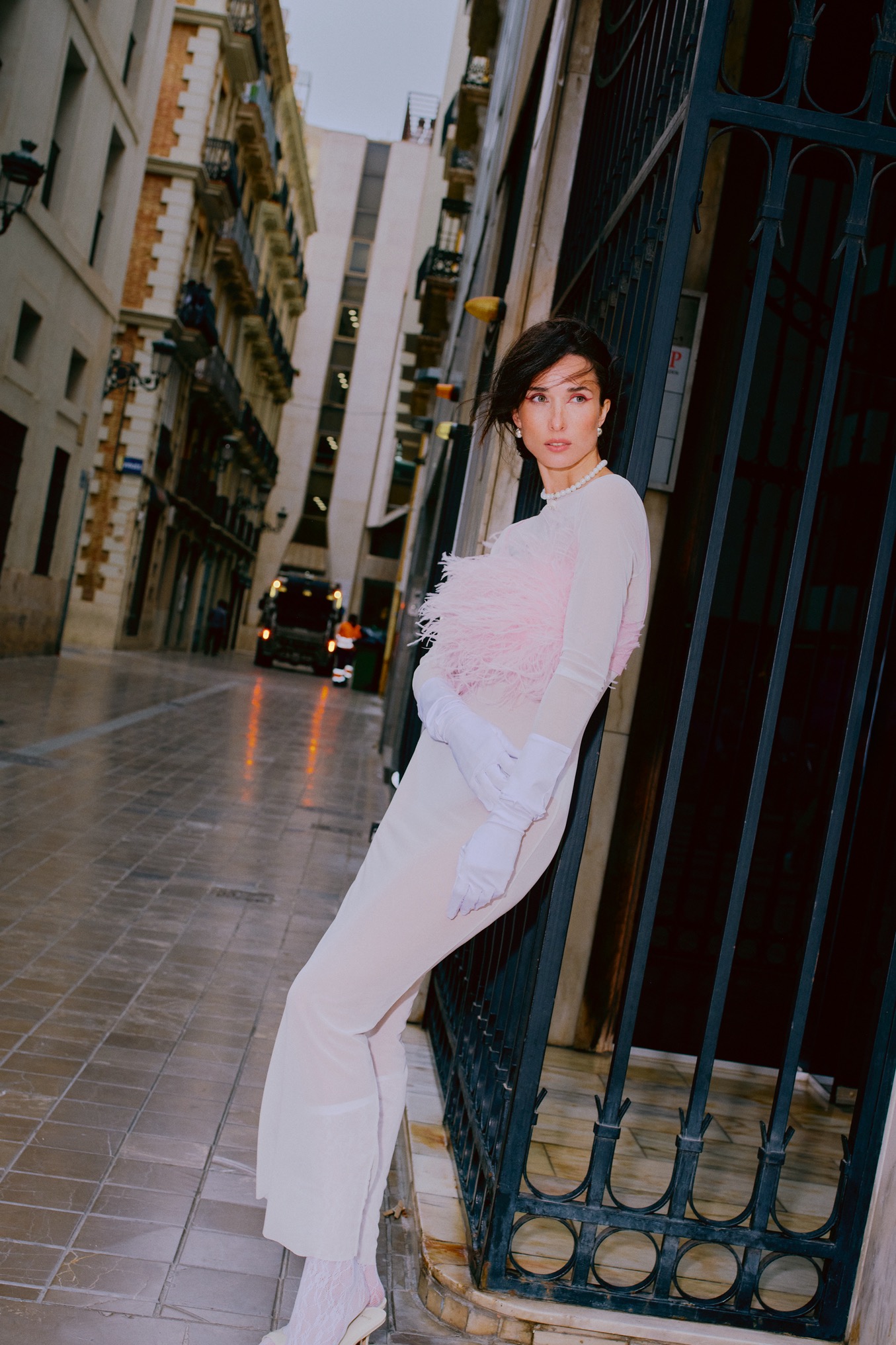 Sesja kobieca w wąskiej uliczce: modelka w białej sukni z piórami i rękawiczkach opiera się o metalową bramę, w tle zabudowa miejska i pojazd komunalny.
