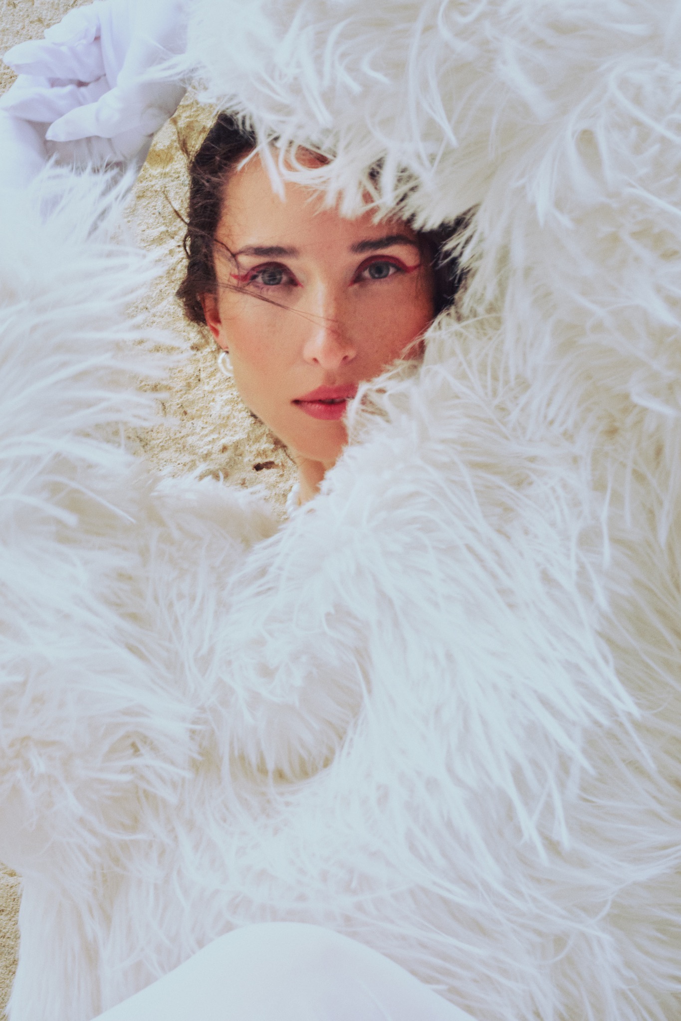 Portret kobiety otulonej białymi piórami na piaskowym tle, z delikatnym makijażem, w jasnym, naturalnym świetle. Ujęcie z góry, podkreślające subtelność i eteryczność.