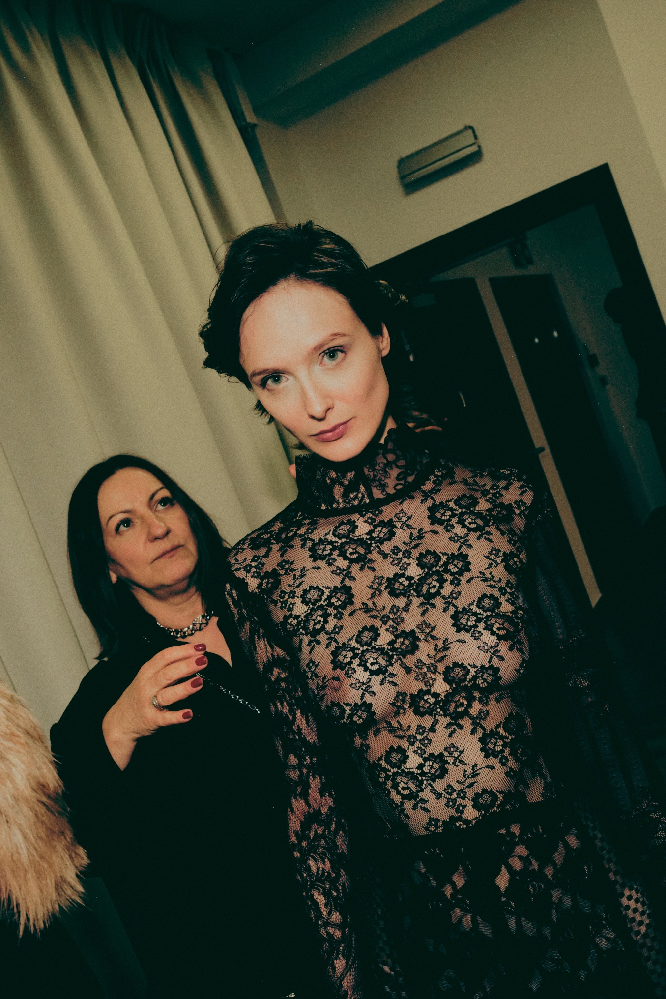 Kobieta w czarnej koronkowej sukience pozuje podczas sesji zdjęciowej. W tle wizażystka poprawia jej włosy. Ujęcie z dołu, ciemne oświetlenie.