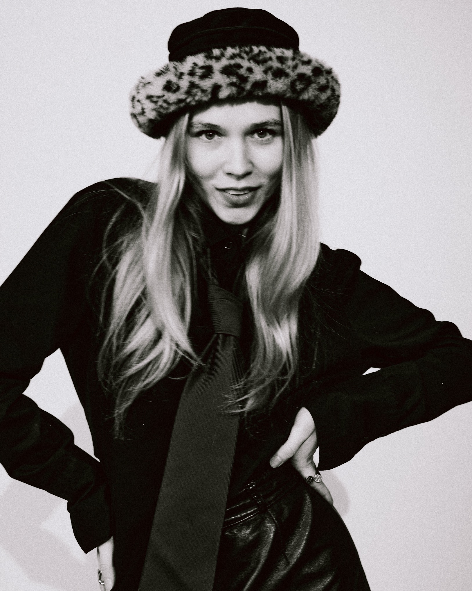 Portret kobiety w czarno-białej tonacji, z kapeluszem w panterkę, ciemną koszulą i krawatem. Uśmiech, dłonie na biodrach, skórzana spódnica.