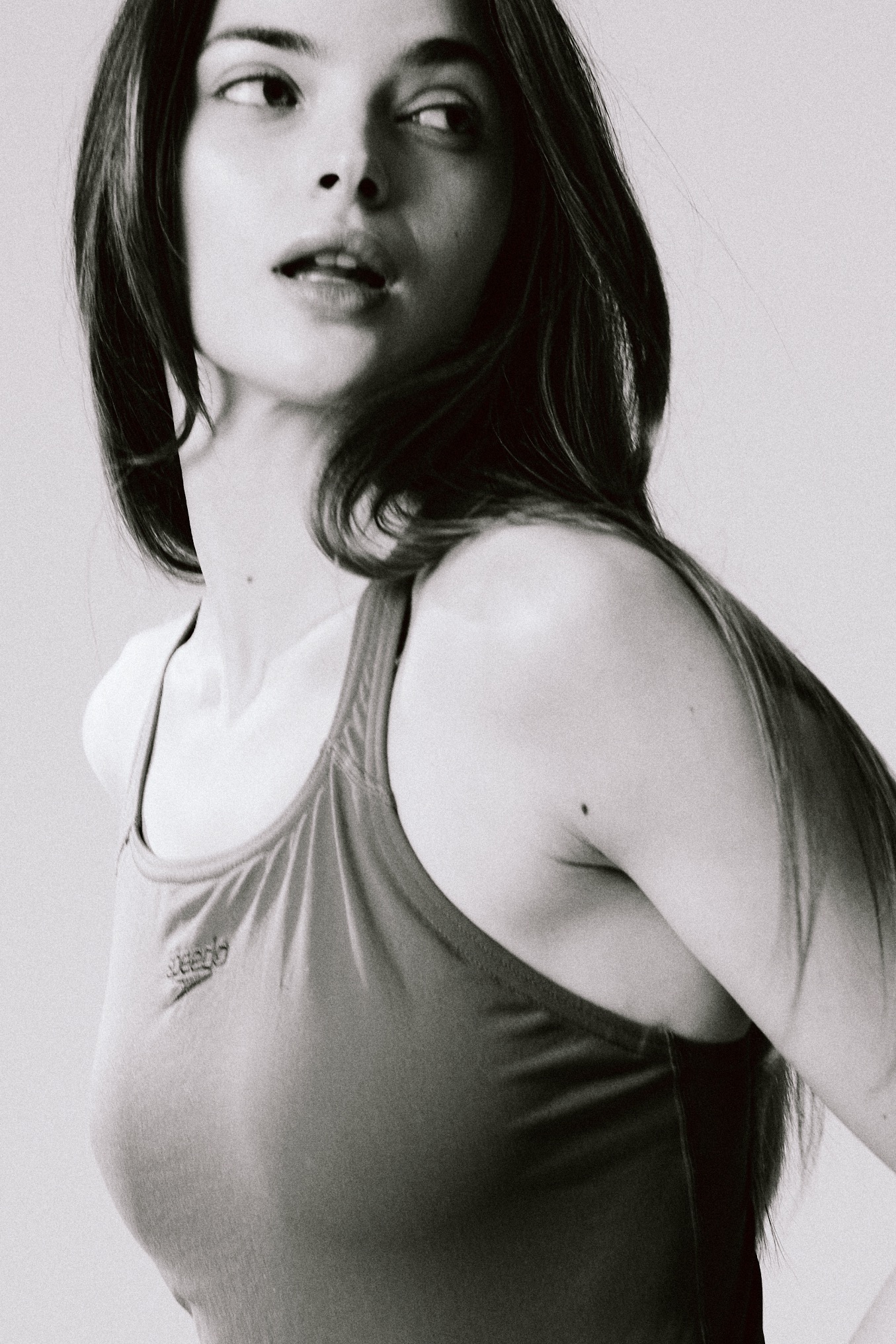 Portret kobiety w sportowym stroju na jasnym tle, skierowanej wzrokiem w lewo, z delikatnym oświetleniem podkreślającym rysy twarzy i fakturę materiału.