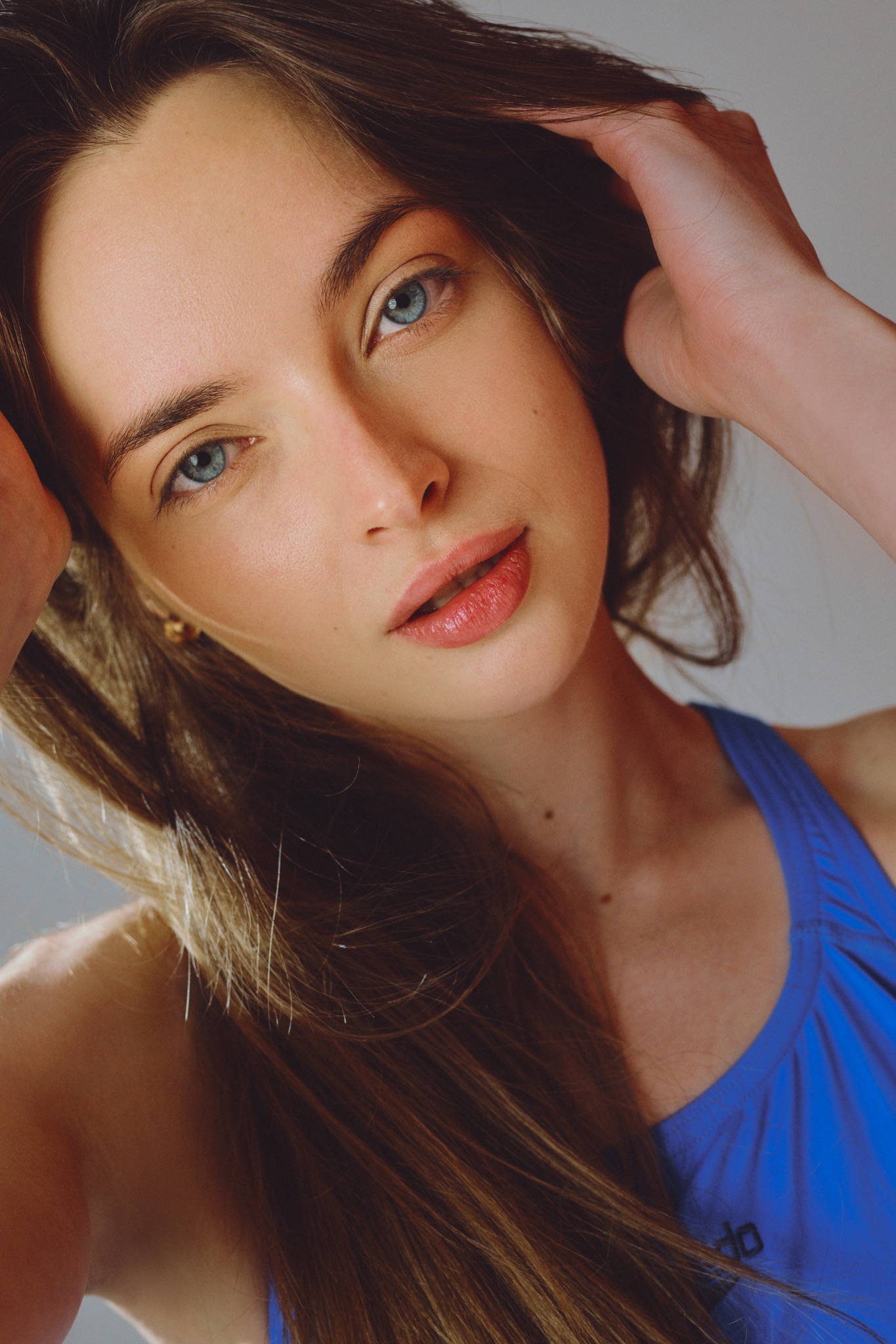Portret młodej kobiety z długimi brązowymi włosami i niebieskimi oczami, ubrana w niebieską koszulkę sportową, pozująca z rękami uniesionymi do włosów.