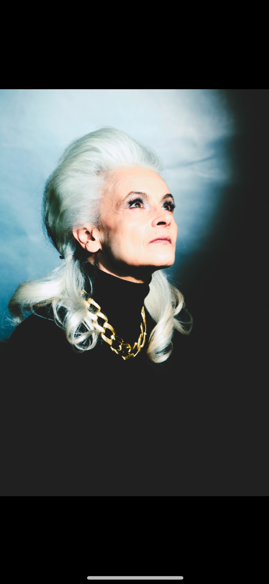 Portret kobiety o siwych włosach, w czarnym golfie i złotym łańcuchu, spoglądającej w górę na tle niebieskiego oświetlenia. Ujęcie od dołu, podkreślające wyraz twarzy.