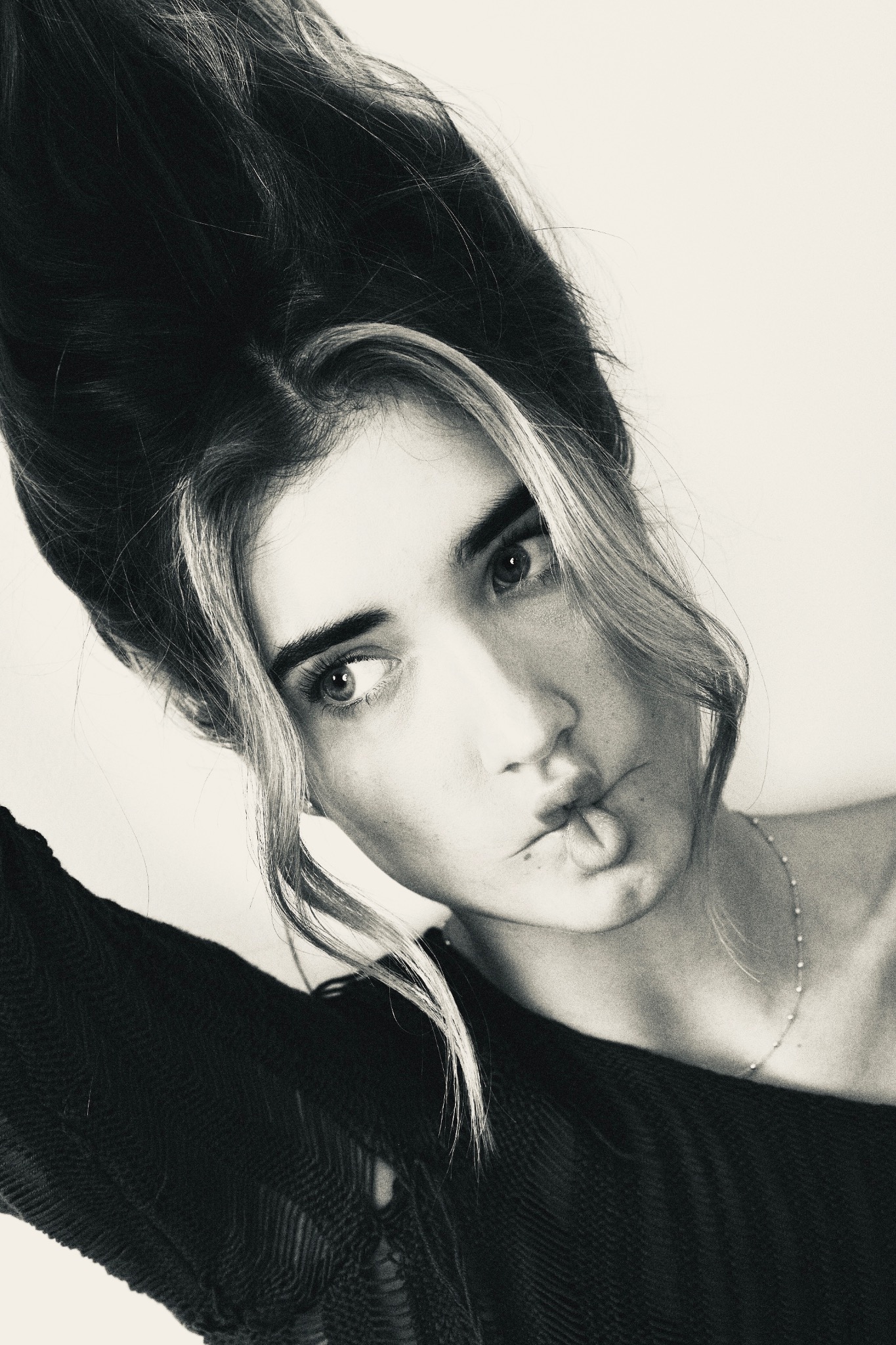 Czarno-biały portret kobiety z uniesionymi włosami, patrzącej w lewo, z delikatnym uśmiechem i subtelną biżuterią na szyi, uchwycony pod nietypowym kątem.