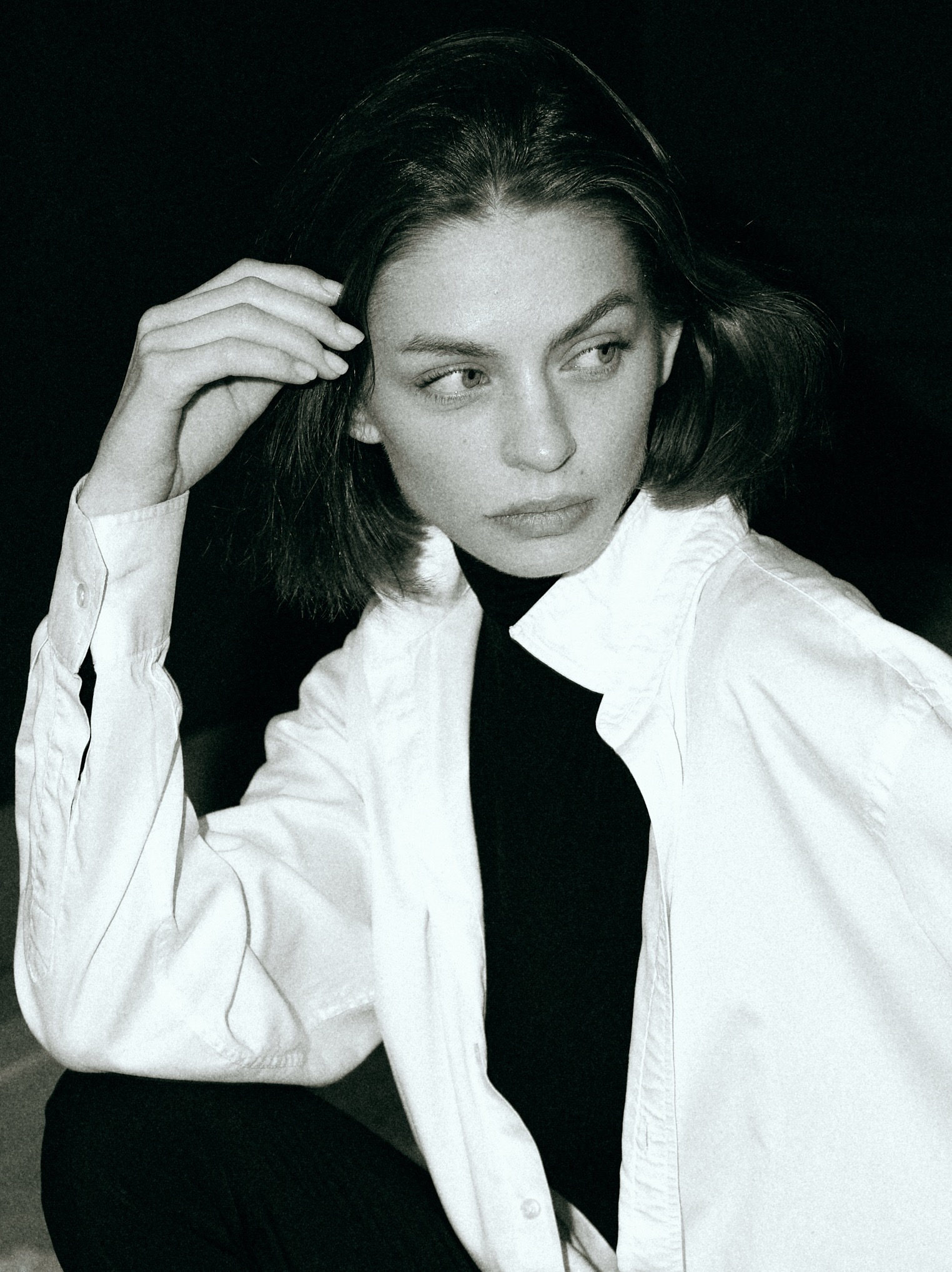 Czarno-biały portret kobiety w białej koszuli i czarnym golfie, z ręką uniesioną do głowy, spoglądającej w bok, na ciemnym tle. Subtelne światło podkreśla rysy twarzy.
