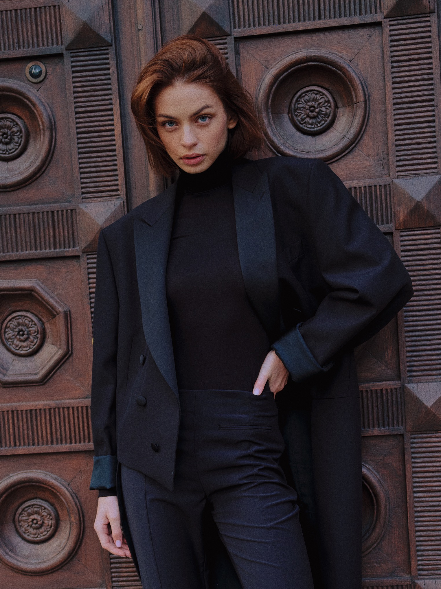 Portret kobiety w czarnym garniturze na tle rzeźbionych, drewnianych drzwi. Ujęcie z bliska, skupione na twarzy i sylwetce modelki. Stonowana kolorystyka, naturalne światło.