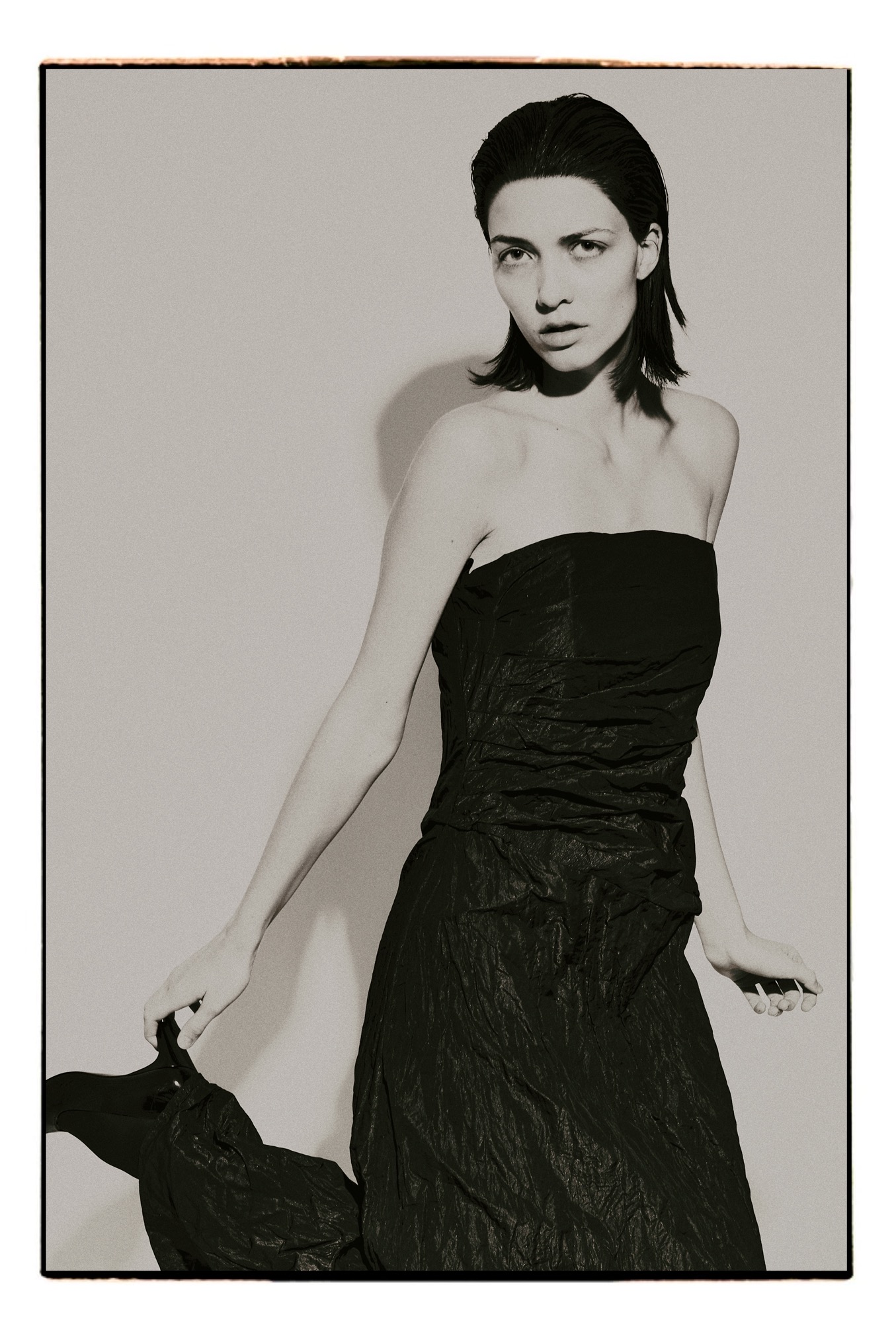 Monochromatyczne ujęcie modelki w czarnej sukni bez ramiączek, z mokrymi włosami, trzymającej w dłoni fragment materiału, na jasnym tle. Minimalistyczny styl, studyjne oświetlenie.